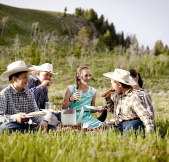 Three children and their parents sit on a picnic rug, drinking juice. Parade Rest Ranch, Montana, USA.