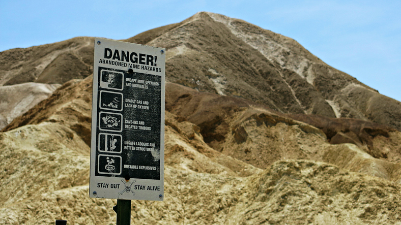 The sign reads: Danger! Abandoned Mine Hazards. Unsafe mine openings and highwalls. Deadly gas and lack of oxygen. Cave-ins and decayed timbers. Unsafe ladders and rotten structures. Unstable structures. Stay out. Stay alive.