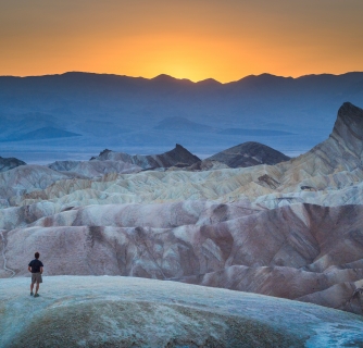 Classic panoramic view of male hiker standing at famous Zabriskie Point viewpoint in beautiful golden evening light at sunset in summer, Death Valley National Park, California, USA