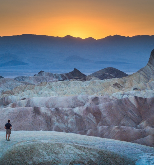 Classic panoramic view of male hiker standing at famous Zabriskie Point viewpoint in beautiful golden evening light at sunset in summer, Death Valley National Park, California, USA