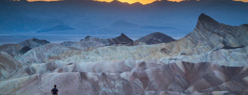 Classic panoramic view of male hiker standing at famous Zabriskie Point viewpoint in beautiful golden evening light at sunset in summer, Death Valley National Park, California, USA