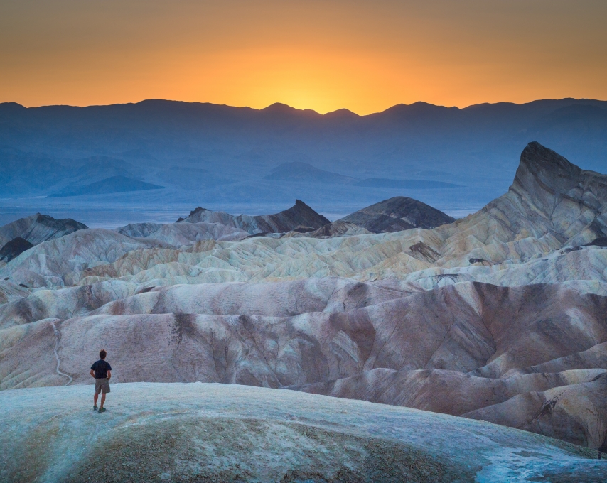 Classic panoramic view of male hiker standing at famous Zabriskie Point viewpoint in beautiful golden evening light at sunset in summer, Death Valley National Park, California, USA