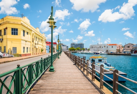 Bright image of wooden promenade at the waterfront of Bridgetown in Barbados. Colorful building against blue sky with white clouds. Boats and yachts in the harbor.
