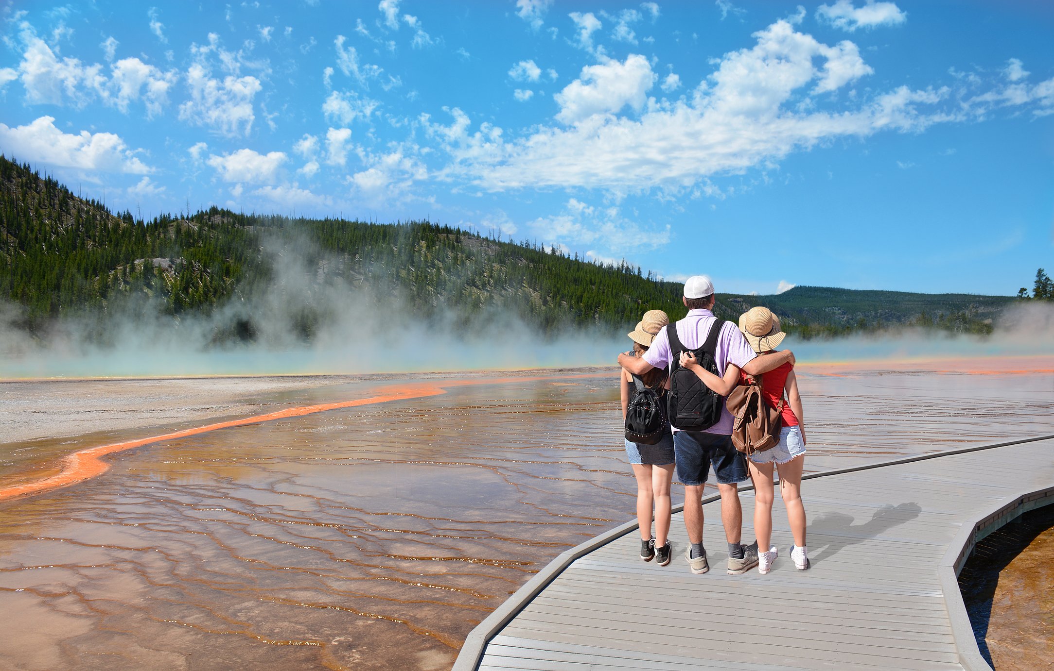 Family hiking on vacation, standing with arms around, , looking at beautiful gazer. Blue sky in the background. Midway Geyser Basin, Grand Prismatic Spring, Yellowstone National Park, USA,